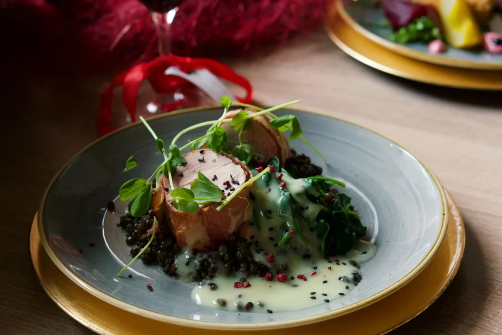 Eleganckie danie restauracyjne: medaliony z polędwiczki wieprzowej owinięte boczkiem, podane na czarnej soczewicy ze szpinakiem, polane jasnym sosem maślanym i udekorowane świeżymi kiełkami groszku.